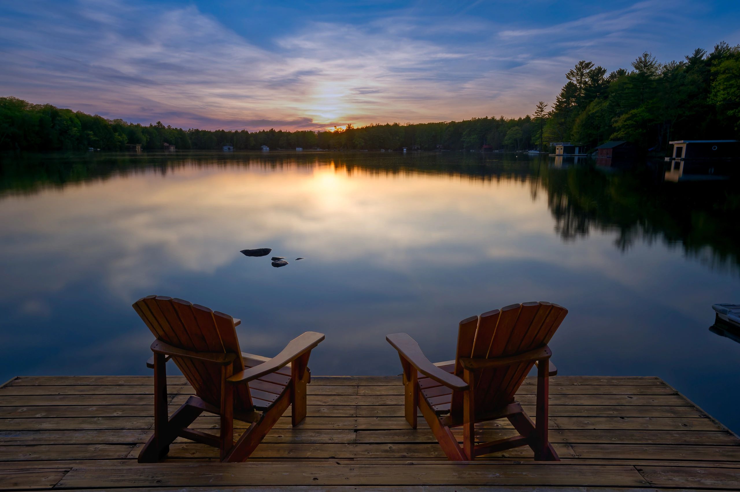 Two Adirondack chairs on a dock overlooking a lake at sunset